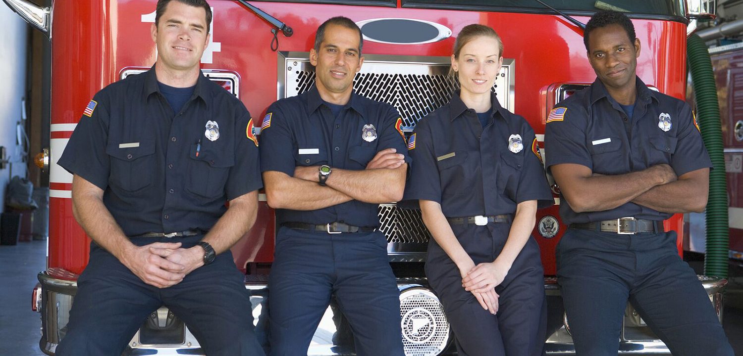 Four firefighters pose in front of their firetruck, smiling for the camera.
