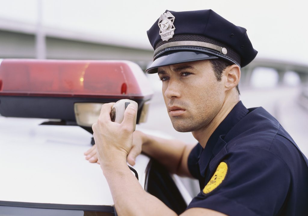 A police officer leans against his cruiser to communicate via radio.