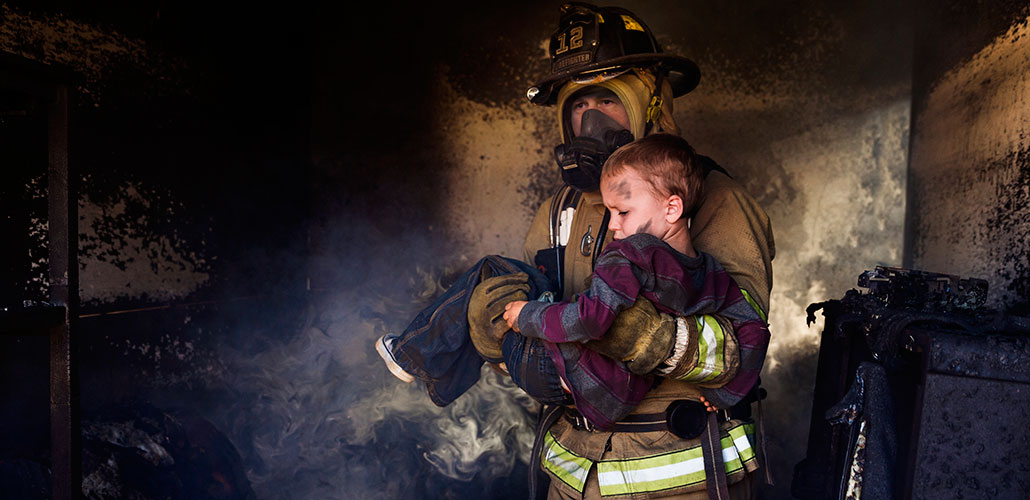A firefighter with a filtered mask carrying a scared and soot-covered child out of a burning building.