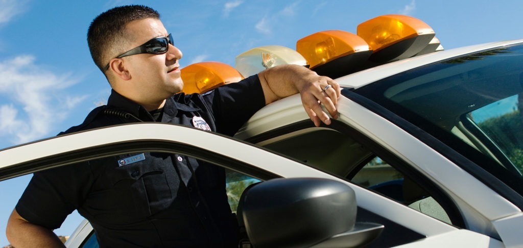 A police officer leans against the inside of his cruiser door looking ahead.
