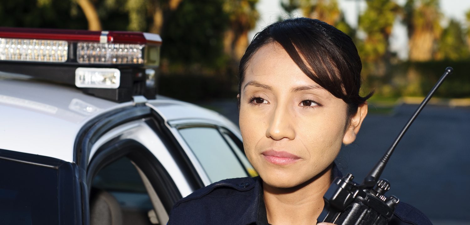 A police officer looks forward whil holding a radio next to her cruiser.