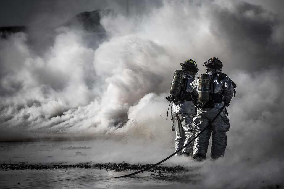 Two well geared firefighters stand at the edge of a large wall of smoke.