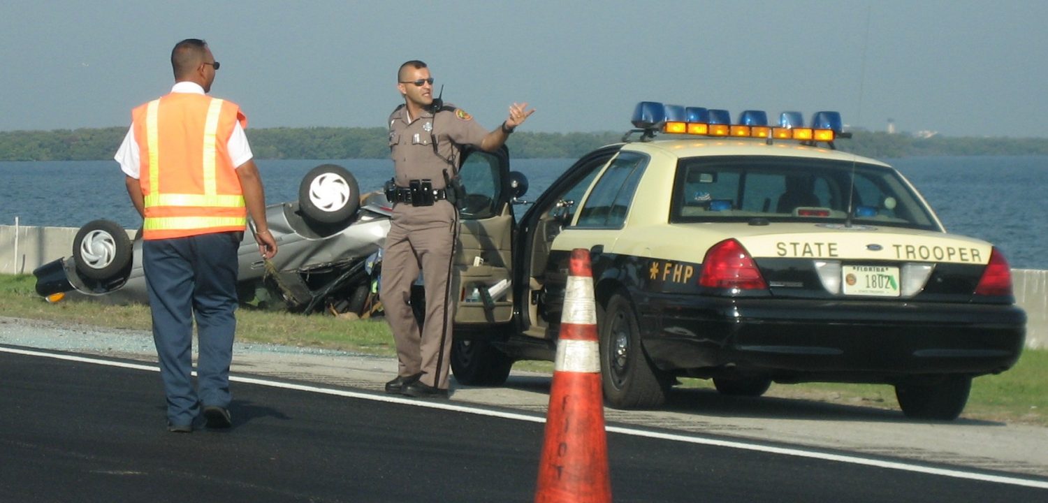 A police officer assists a man in reflective clothing in coordinating a vehicle incident off the side of a roadway.