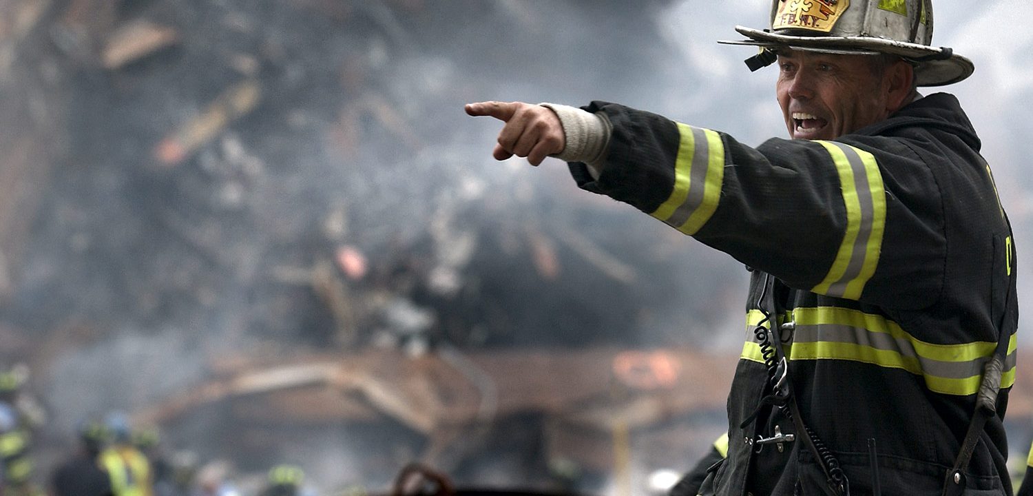 A firefighter points while standing in front of a pile of burning rubble and other workers.