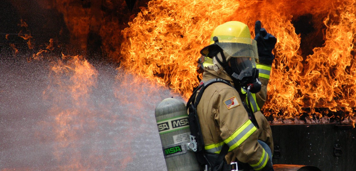 A firefighter signals to another person whilst water sprays a roaring flame during an exercise.