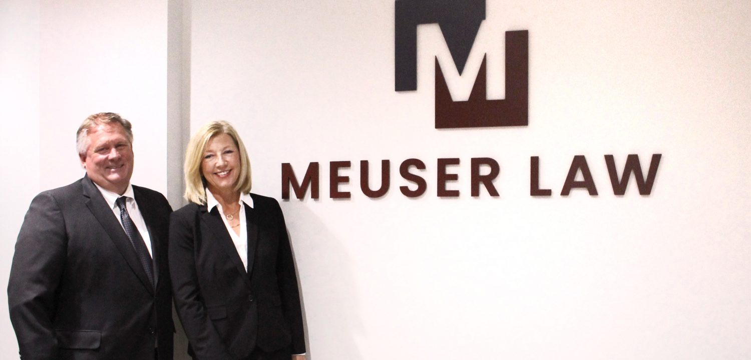 Ron and Nancy stand together and smile for the camera next to a Meuser Law logo on a wall.