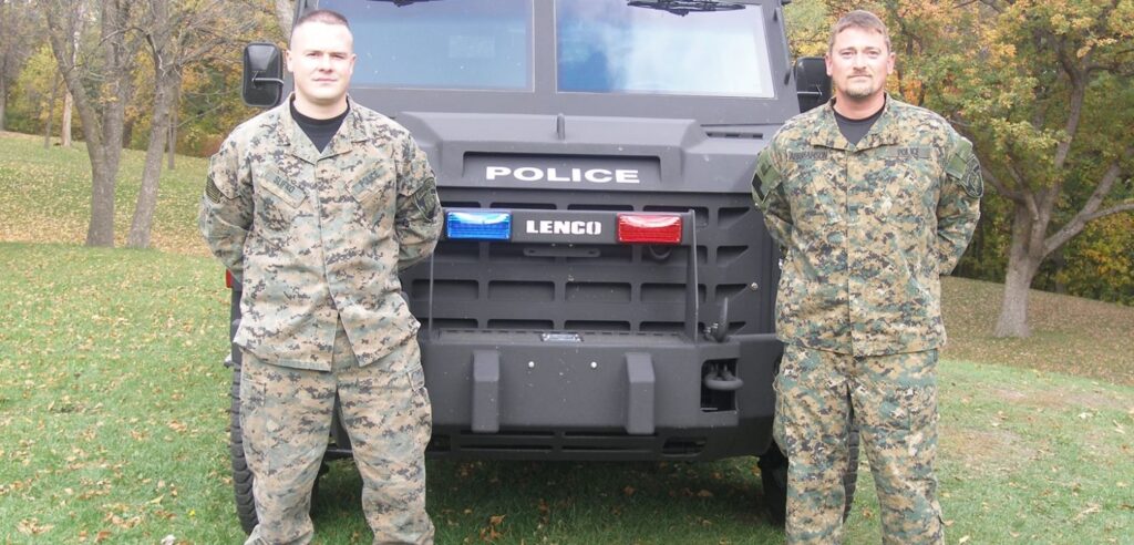 Two men in military fatiques pose in front of an armored vehicle.