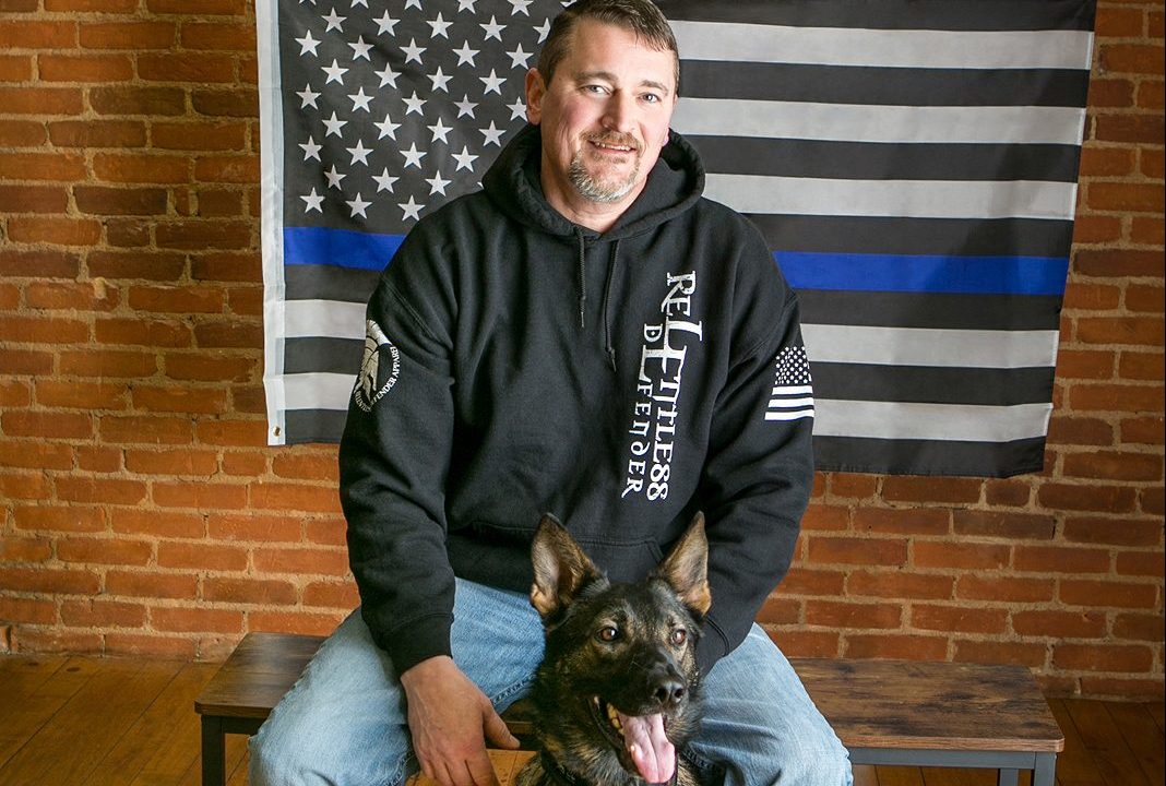 A man sits on a bench with his dog in front of a thin blue line American flag.
