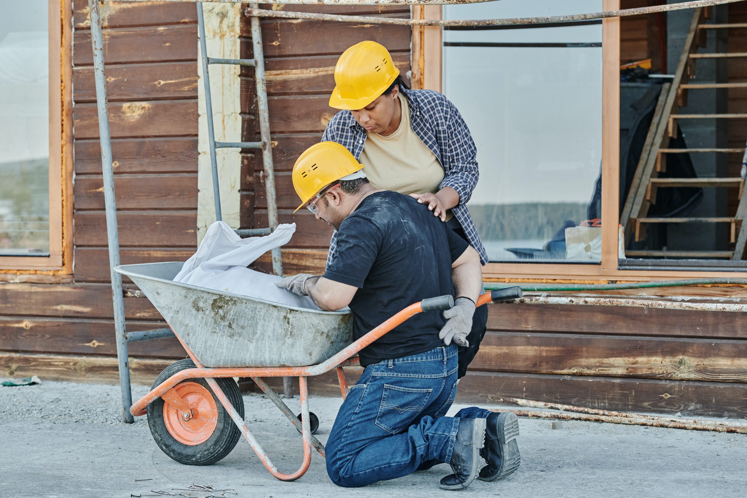 A worker kneels down next to his wheel barrow and touches his pained lower back while another worker attempts to help him.